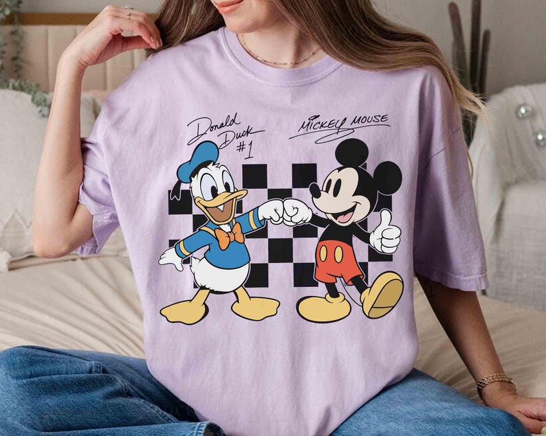 May include: A light purple t-shirt featuring Donald Duck and Mickey Mouse. Donald Duck is wearing a blue sailor suit and Mickey Mouse is wearing red shorts. The text "Donald Duck #1" and "Mickey Mouse" are above the characters.