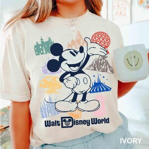 Może przedstawiać: Kremowa koszulka z grafiką Myszki Miki i r&oacute;żnymi zabytkami Walt Disney World. Koszulka zawiera napis "Walt Disney World". Widoczny jest r&oacute;wnież kubek z uśmiechniętą buzią.