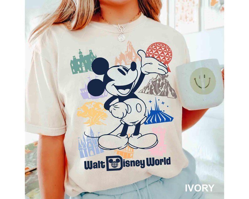 Może przedstawiać: Kremowa koszulka z grafiką Myszki Miki i r&oacute;żnymi zabytkami Walt Disney World. Koszulka zawiera napis "Walt Disney World". Widoczny jest r&oacute;wnież kubek z uśmiechniętą buzią.