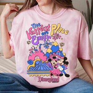 K&ouml;nnte beinhalten: Ein hellrosa T-Shirt mit dem Text "The Happiest Place on Earth" in farbenfrohen, verspielten Buchstaben. Die Grafik zeigt Micky und Minnie Maus vor einer stilisierten Disneyland-Szene.