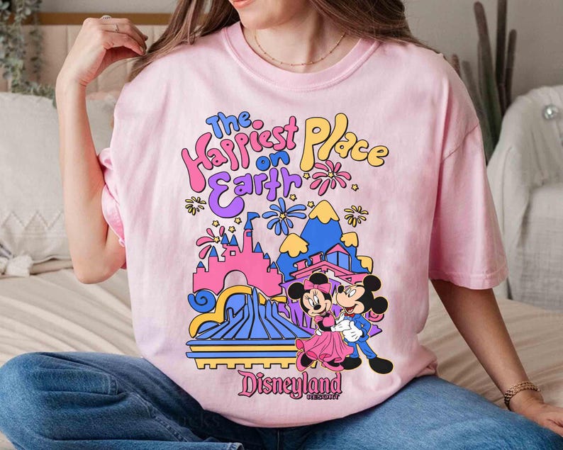 K&ouml;nnte beinhalten: Ein hellrosa T-Shirt mit dem Text "The Happiest Place on Earth" in farbenfrohen, verspielten Buchstaben. Die Grafik zeigt Micky und Minnie Maus vor einer stilisierten Disneyland-Szene.