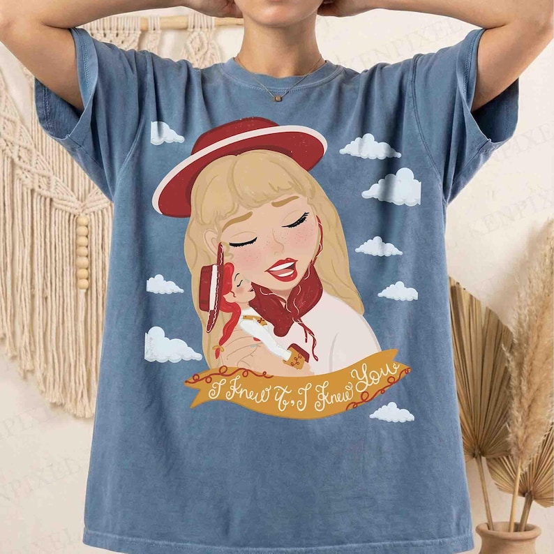 May include: A blue short-sleeve t-shirt featuring a graphic illustration of a blonde character wearing a red hat and holding a small doll. The design includes white clouds and a yellow banner with the text I Knew It, I Knew You printed in brown.