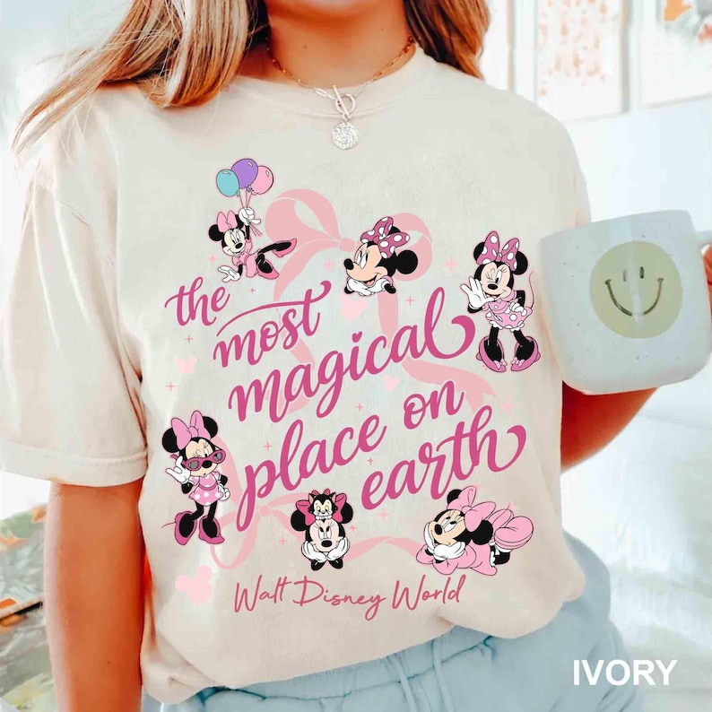 K&ouml;nnte beinhalten: Elfenbeinfarbenes T-Shirt mit Minnie-Maus-Illustrationen und dem pinkfarbenen Text "the most magical place on earth". Minnie Mouse wird in verschiedenen Posen gezeigt, mit rosa Schleifen und Kleidern. Das Shirt enth&auml;lt auch den Text "Walt Disney World".
