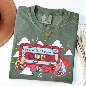May include: An olive green short-sleeve t-shirt featuring a graphic of a red cassette tape with the text "I KNEW IT, I KNEW YOU" and "TS". The design includes a yellow sheriff star with the number 13, white clouds, stars, musical notes, and a cowboy boot with a red hat.