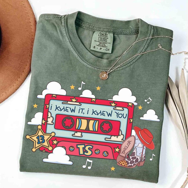 May include: An olive green short-sleeve t-shirt featuring a graphic of a red cassette tape with the text "I KNEW IT, I KNEW YOU" and "TS". The design includes a yellow sheriff star with the number 13, white clouds, stars, musical notes, and a cowboy boot with a red hat.