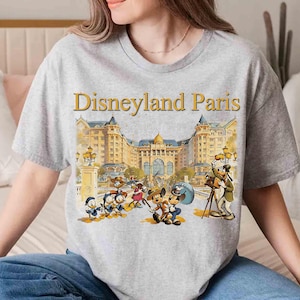 May include: A gray t-shirt featuring the words "Disneyland Paris" above a colorful illustration of Disney characters in front of a grand building. The design includes Mickey Mouse, Minnie Mouse, Donald Duck, and Goofy.