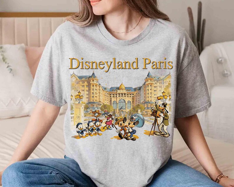 May include: A gray t-shirt featuring the words "Disneyland Paris" above a colorful illustration of Disney characters in front of a grand building. The design includes Mickey Mouse, Minnie Mouse, Donald Duck, and Goofy.