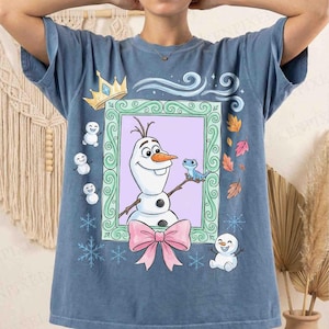 May include: A blue short-sleeve t-shirt featuring a central graphic of a snowman and a small blue lizard inside a green ornate frame. The design includes a pink bow, small snowmen, snowflakes, a gold crown, and autumn leaves around the frame.