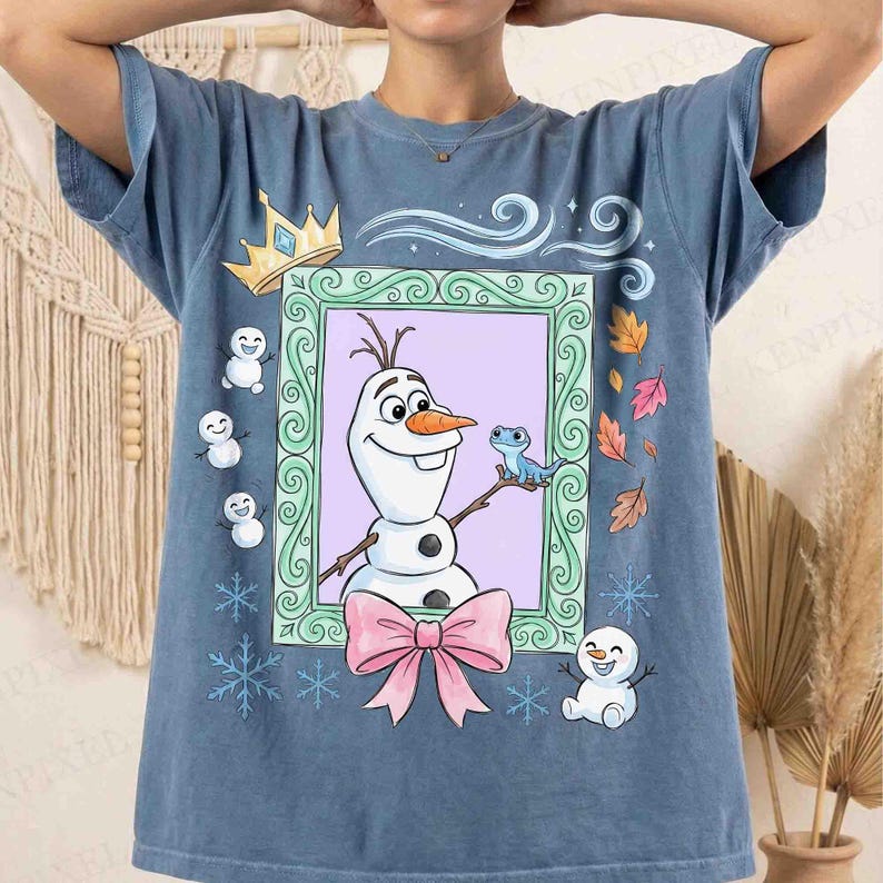 May include: A blue short-sleeve t-shirt featuring a central graphic of a snowman and a small blue lizard inside a green ornate frame. The design includes a pink bow, small snowmen, snowflakes, a gold crown, and autumn leaves around the frame.
