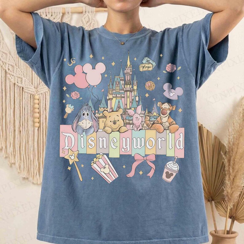 May include: Blue t-shirt featuring a colorful Disneyworld graphic. The design includes Disney characters like Winnie the Pooh, Eeyore, and Tigger, along with a castle, balloons, and the word "Disneyworld".
