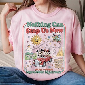 Peut inclure: T-shirt rose clair avec un motif color&eacute;. Le motif comprend le texte "Nothing Can Stop Us Now", un soleil souriant, un train et une voiture rouge avec Mickey et Minnie Mouse. Le t-shirt est de style col rond.