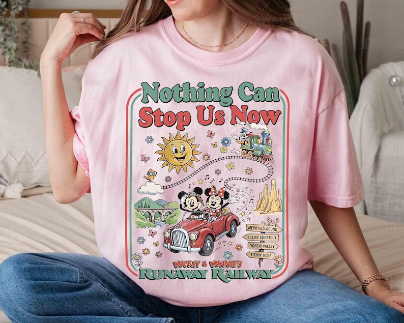 Peut inclure: T-shirt rose clair avec un motif color&eacute;. Le motif comprend le texte "Nothing Can Stop Us Now", un soleil souriant, un train et une voiture rouge avec Mickey et Minnie Mouse. Le t-shirt est de style col rond.