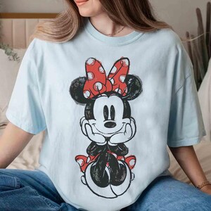 May include: Pale blue t-shirt with a Minnie Mouse illustration. The design features Minnie with a red polka dot bow and a black and white outline. The shirt has short sleeves and a relaxed fit.