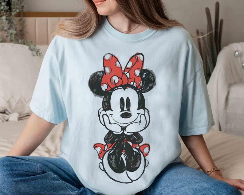 May include: Pale blue t-shirt with a Minnie Mouse illustration. The design features Minnie with a red polka dot bow and a black and white outline. The shirt has short sleeves and a relaxed fit.
