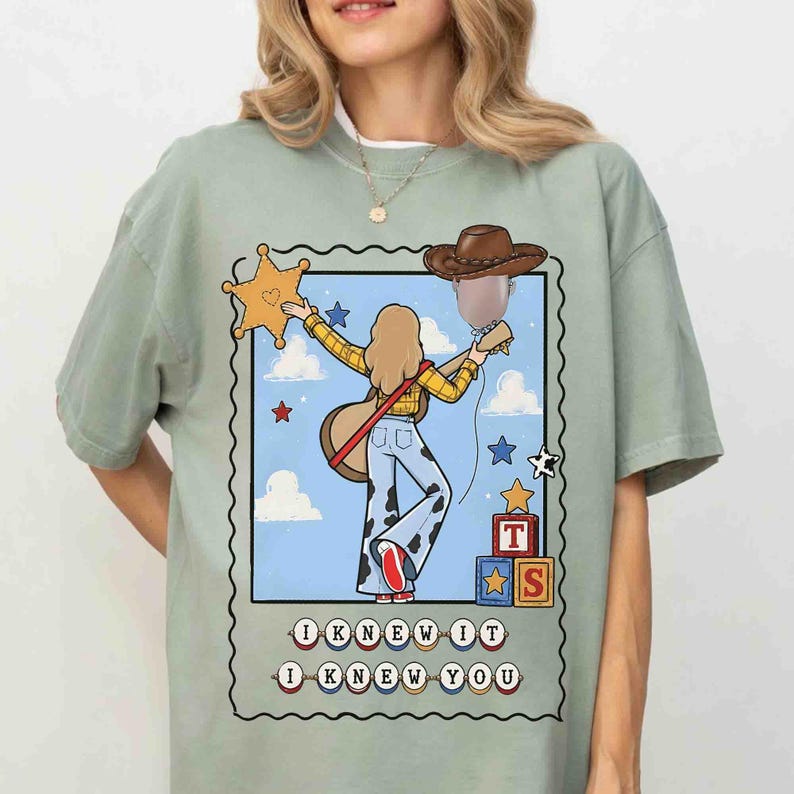 May include: A sage green t-shirt featuring a graphic illustration of a person with long hair wearing a yellow plaid shirt and cow-print bell-bottoms, holding a guitar. The design includes a sheriff badge, a cowboy hat, and blocks with the letters T and S, with the text I KNEW YOU below.