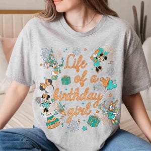 May include: A gray t-shirt with the text "Life of a birthday girl" in orange script. The shirt features cartoon characters, gifts, fireworks, and a birthday cake design. The characters include Minnie Mouse, Mickey Mouse, and Daisy Duck.