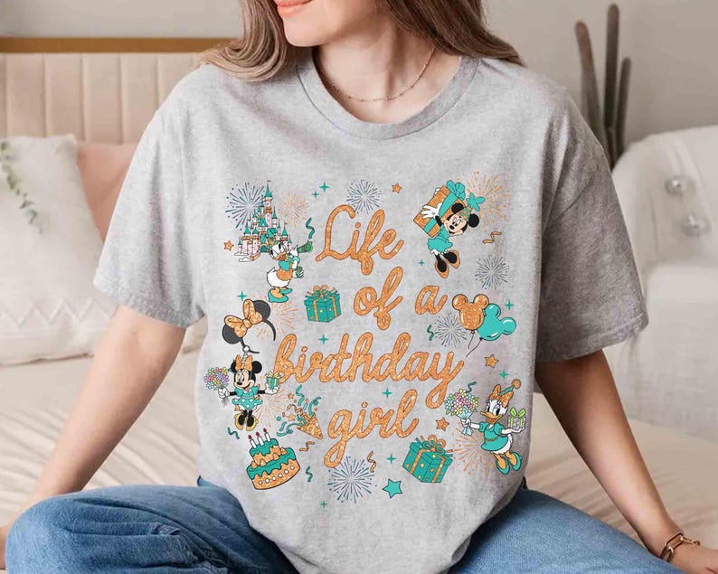 May include: A gray t-shirt with the text "Life of a birthday girl" in orange script. The shirt features cartoon characters, gifts, fireworks, and a birthday cake design. The characters include Minnie Mouse, Mickey Mouse, and Daisy Duck.