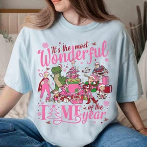 May include: Light blue t-shirt with a pink graphic that reads "It's the most Wonderful Time of the Year." The graphic features cartoon characters, a Christmas tree, and wrapped gifts. The shirt is a relaxed fit.