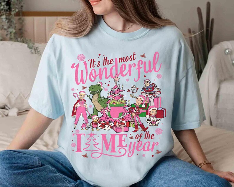 May include: Light blue t-shirt with a pink graphic that reads "It's the most Wonderful Time of the Year." The graphic features cartoon characters, a Christmas tree, and wrapped gifts. The shirt is a relaxed fit.