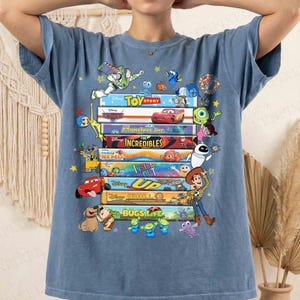 May include: Blue t-shirt featuring a stack of colorful movie titles and characters from Disney and Pixar films, including "Toy Story," "Cars," "Monsters, Inc.," "The Incredibles," "Finding Nemo," "Inside Out," "Up," "WALL-E," and "A Bug's Life." The design includes various animated characters.