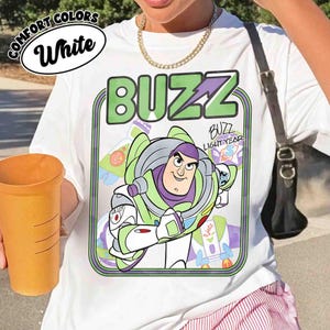 May include: A white t-shirt featuring a graphic of the character Buzz Lightyear in his signature green, purple, and white space suit. The design includes the text BUZZ in large green letters at the top and the name Buzz Lightyear in script.