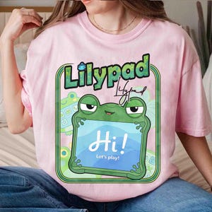 May include: A light pink t-shirt featuring a graphic of a green frog holding a blue tablet screen that displays the text "Hi! Let's play!". Above the frog, the word "Lilypad" is printed in a bold, green, textured font with a small frog icon.