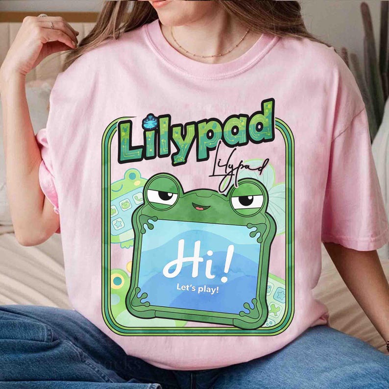 May include: A light pink t-shirt featuring a graphic of a green frog holding a blue tablet screen that displays the text "Hi! Let's play!". Above the frog, the word "Lilypad" is printed in a bold, green, textured font with a small frog icon.