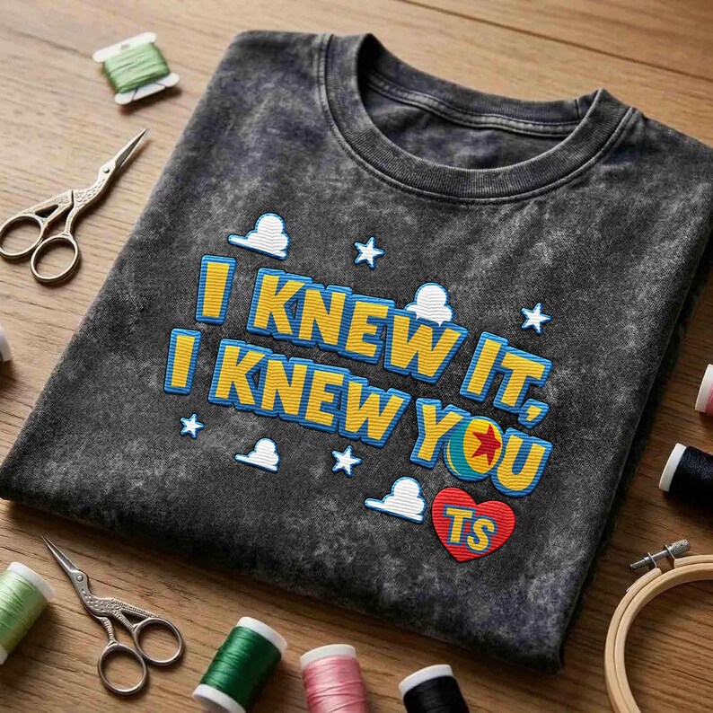 May include: A charcoal gray acid-wash t-shirt featuring the embroidered yellow text I KNEW IT, I KNEW YOU with blue outlines, white cloud and star icons, a colorful ball graphic, and a red heart with the letters TS embroidered on the front.