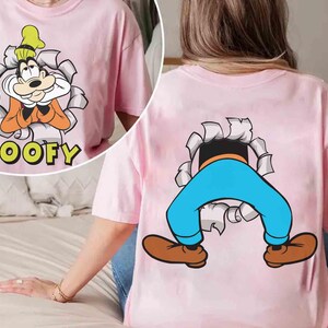 May include: Pink t-shirt featuring a cartoon image of Goofy bursting through a torn paper effect. The front shows Goofy's face with the word "GOOFY" below. The back displays Goofy's legs and feet emerging from a similar tear.