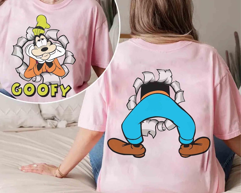May include: Pink t-shirt featuring a cartoon image of Goofy bursting through a torn paper effect. The front shows Goofy's face with the word "GOOFY" below. The back displays Goofy's legs and feet emerging from a similar tear.