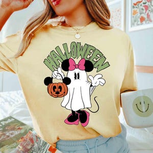 May include: Yellow t-shirt featuring a Halloween-themed Minnie Mouse design. The graphic shows Minnie as a ghost holding a jack-o'-lantern. The word "HALLOWEEN" is arched above the character in green letters.