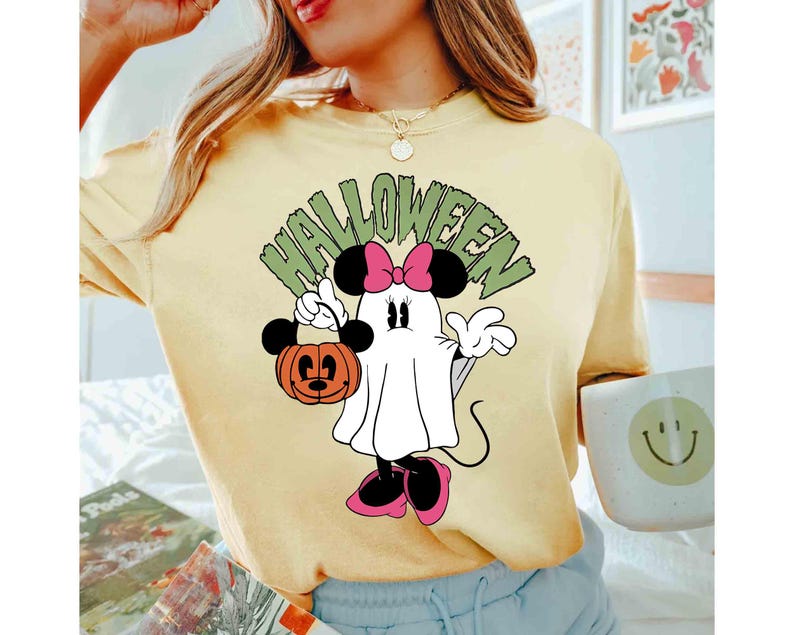 May include: Yellow t-shirt featuring a Halloween-themed Minnie Mouse design. The graphic shows Minnie as a ghost holding a jack-o'-lantern. The word "HALLOWEEN" is arched above the character in green letters.