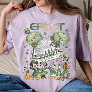 May include: A lilac t-shirt with the word "EPCOT" in green and gold. The design features Mickey and friends in green St. Patrick's Day outfits, a dragon, and the EPCOT sphere. The shirt has a loose fit.