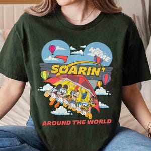 May include: Dark green t-shirt featuring a colorful graphic of Mickey Mouse, Goofy, and Donald Duck hang gliding. The design includes hot air balloons, clouds, and the words "SOARIN'" and "AROUND THE WORLD". The shirt has a crew neck and short sleeves.