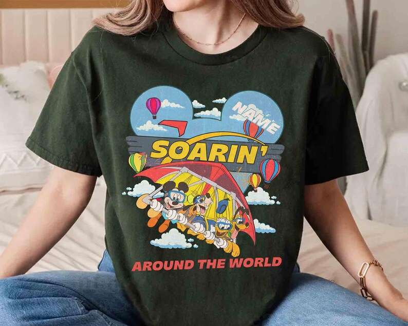 May include: Dark green t-shirt featuring a colorful graphic of Mickey Mouse, Goofy, and Donald Duck hang gliding. The design includes hot air balloons, clouds, and the words "SOARIN'" and "AROUND THE WORLD". The shirt has a crew neck and short sleeves.