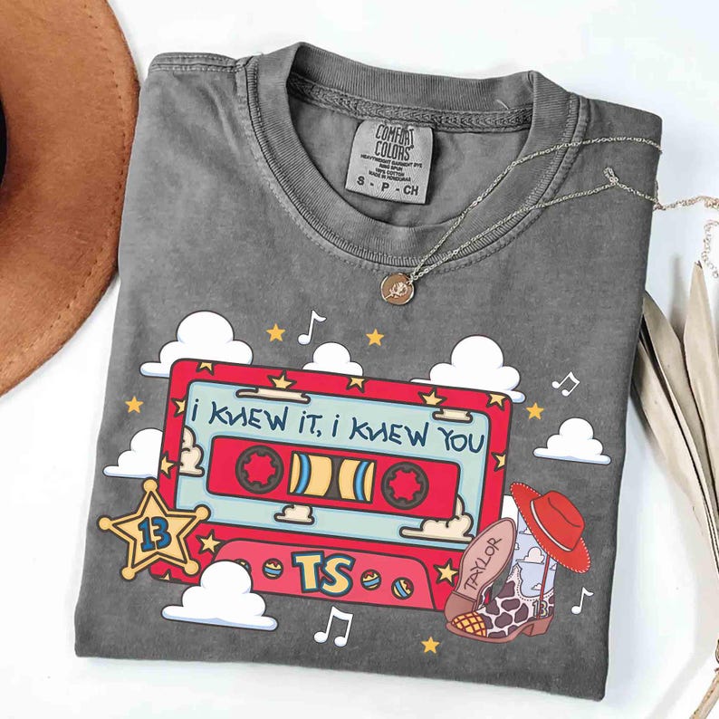 May include: A charcoal grey short-sleeve t-shirt featuring a graphic of a red cassette tape with the text "I KNEW IT, I KNEW YOU" and "TS". The design includes a yellow sheriff star with the number 13, white clouds, musical notes, and a cowboy boot.