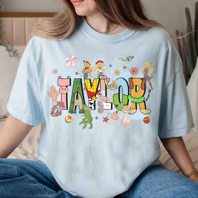 May include: A light blue t-shirt featuring the name TAYLOR in large, colorful block letters. The design is decorated with various cartoon characters, small stars, flowers, a bow, and a rocket ship scattered around the text in a playful arrangement.