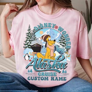 May include: Pink t-shirt featuring a graphic of Pluto in winter attire, with the words "Disney Wonder Alaska Cruise Custom Name." The design includes a cruise ship, mountains, and a penguin.