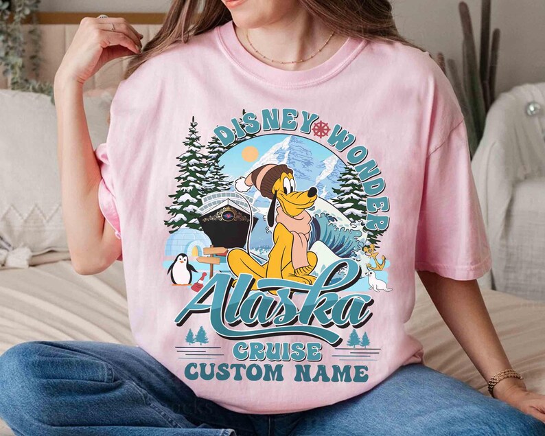 May include: Pink t-shirt featuring a graphic of Pluto in winter attire, with the words "Disney Wonder Alaska Cruise Custom Name." The design includes a cruise ship, mountains, and a penguin.