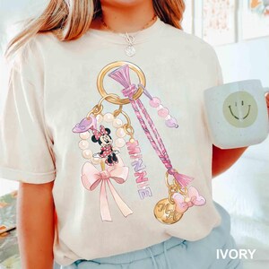 Op de afbeelding: Ivoor kleurig T-shirt met een Minnie Mouse sleutelhanger ontwerp. Het ontwerp bevat een gouden ring, roze strik, parels en het woord "MINNIE". Een witte mok met een smiley is ook zichtbaar.