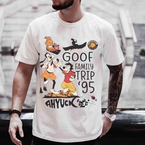 Pode incluir: Camiseta branca com um gr&aacute;fico de desenho animado colorido. O gr&aacute;fico inclui o texto "GOOF FAMILY TRIP '95" e "AHYUCK", juntamente com personagens de desenhos animados e imagens de um barco, um carro e uma cena de montanha.