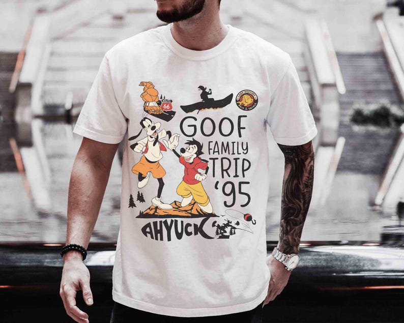 Pode incluir: Camiseta branca com um gr&aacute;fico de desenho animado colorido. O gr&aacute;fico inclui o texto "GOOF FAMILY TRIP '95" e "AHYUCK", juntamente com personagens de desenhos animados e imagens de um barco, um carro e uma cena de montanha.
