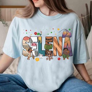 Puede incluir: Camiseta azul claro con el nombre "SELNA" en letras grandes y coloridas. Cada letra est&aacute; decorada con personajes de dibujos animados y elementos tropicales. La camiseta la lleva una persona.