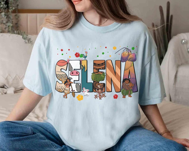 Puede incluir: Camiseta azul claro con el nombre "SELNA" en letras grandes y coloridas. Cada letra est&aacute; decorada con personajes de dibujos animados y elementos tropicales. La camiseta la lleva una persona.