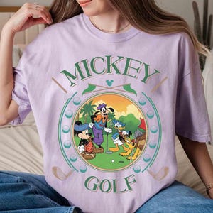 May include: A light purple t-shirt featuring a circular graphic of Mickey Mouse, Donald Duck, Goofy, and Pluto playing golf. The words "Mickey Golf" are printed above and below the graphic in green and white.