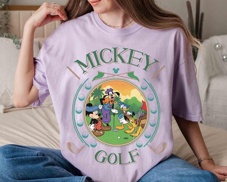 May include: A light purple t-shirt featuring a circular graphic of Mickey Mouse, Donald Duck, Goofy, and Pluto playing golf. The words "Mickey Golf" are printed above and below the graphic in green and white.