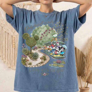 May include: Blue t-shirt featuring a colorful graphic of Disney characters, including Mickey and Minnie Mouse, in a whimsical garden setting. The shirt has the text "Only the will is truly living with the god".