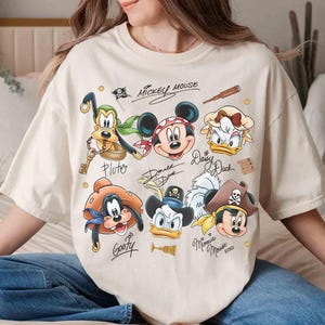 May include: Cream-colored t-shirt featuring cartoon pirate versions of Mickey Mouse, Minnie Mouse, Donald Duck, Daisy Duck, Goofy, and Pluto. The shirt includes the text "Mickey Mouse" and character signatures. The design has a vintage, distressed look.