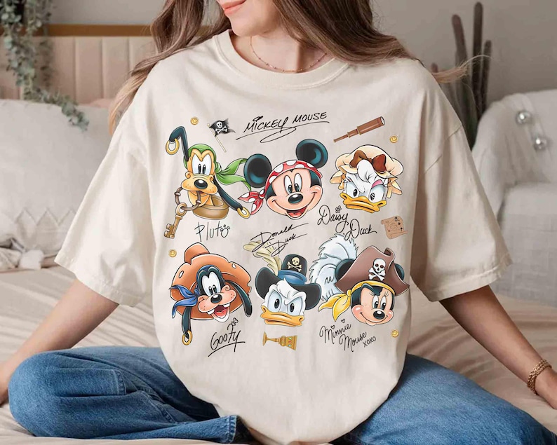May include: Cream-colored t-shirt featuring cartoon pirate versions of Mickey Mouse, Minnie Mouse, Donald Duck, Daisy Duck, Goofy, and Pluto. The shirt includes the text "Mickey Mouse" and character signatures. The design has a vintage, distressed look.