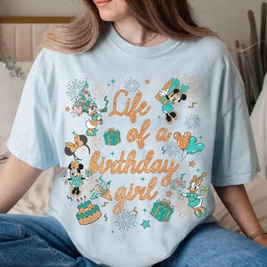 May include: Light blue t-shirt with the text "Life of a birthday girl" in orange script. The shirt features cartoon characters, including Mickey and Minnie Mouse, and Daisy Duck, along with birthday-themed graphics like fireworks, gifts, and a cake.